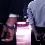 Two men in handcuffs with police lights in the background