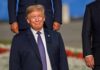 Trump – NYC Deal Talk Teases Big Build smiling man in a navy suit at an outdoor event