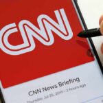 CNN Booted; Who Controls The Tape? CNN logo on a smartphone screen with a hand holding a stylus