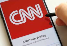 CNN Booted; Who Controls The Tape? CNN logo on a smartphone screen with a hand holding a stylus