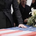 Culture War Erupts Over One Scarf Mourner rests hand on flag-draped casket with flower arrangement