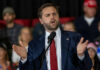 Speculative Smear Explodes Onstage Man in suit speaking at a podium