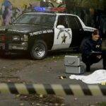 City Employee Linked To Cop Ambush Crime scene with police car and two investigators by a covered body