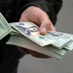 Six-Figure Checks Spark California Outrage A hand holding a stack of hundred dollar bills with one bill prominently displayed